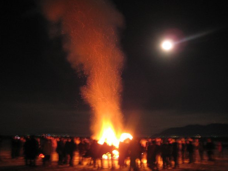 Large bonfire at night under a full moon with a crowd gathered